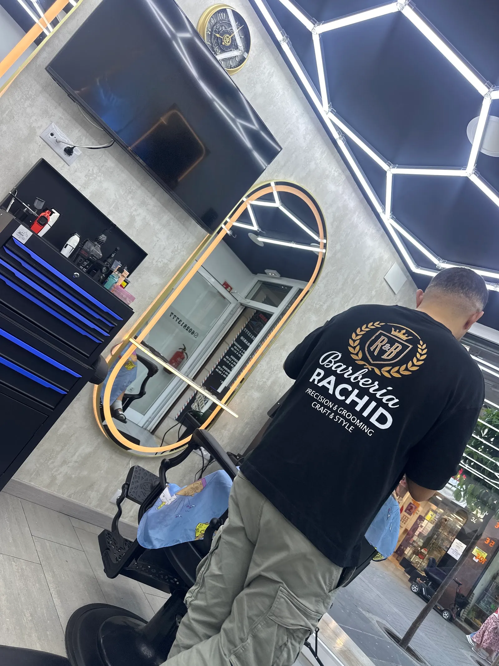 Barbero de espaldas con la camiseta de Barbería Rachid, frente a un espejo dorado